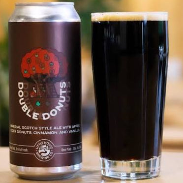 A can of Double Donuts Non-Alcoholic Scotcht Style Ale next to a tall glass filled with dark beer, set on a wooden surface with a softly blurred indoor background.